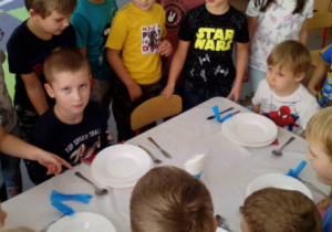 Grupa dzieci zebrana wokół stolika, który został nakryty do posiłku, ocenia poprawność wykonania zadania swych kolegów.
